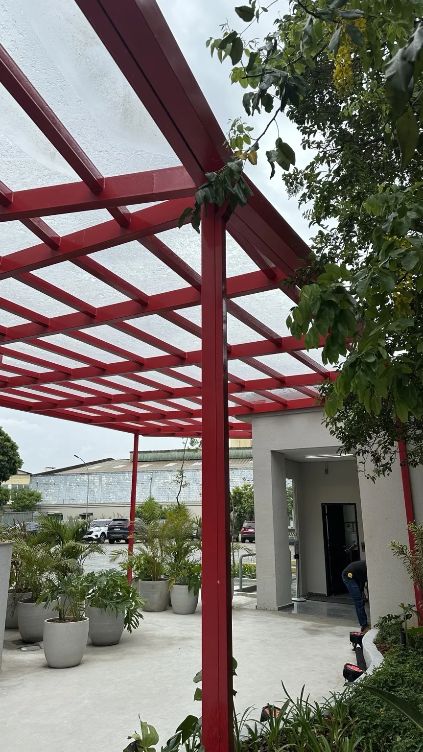 Cobertura transparente com estrutura metálica vermelha sobre área ajardinada